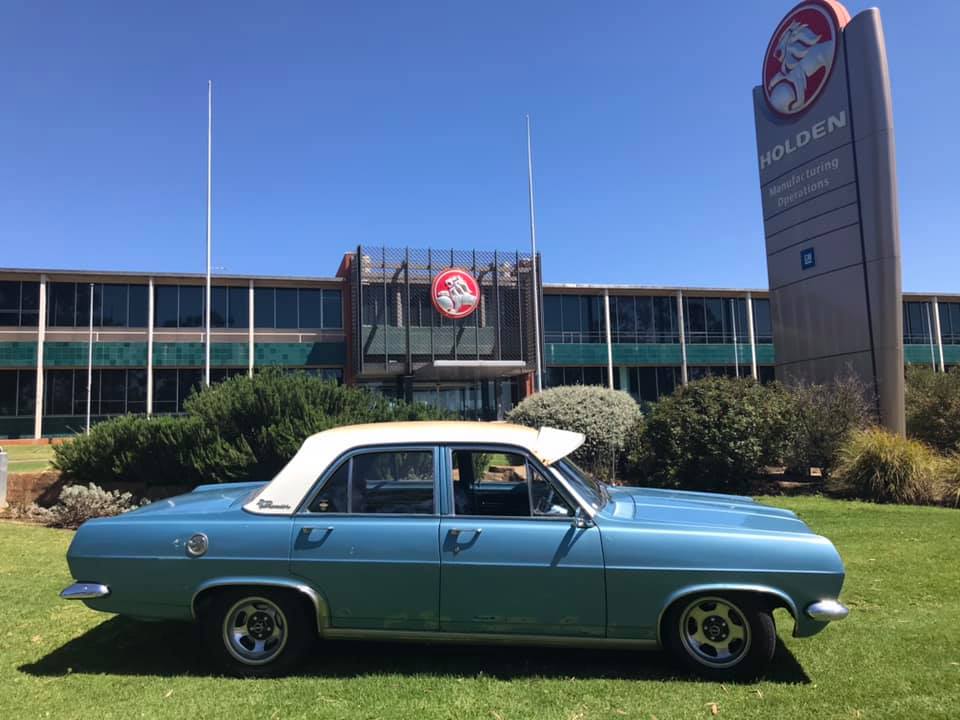 An Holden HR Premier Sedan is parked in front of the Holden offices at Elizabeth, South Australia. The car’s roof is white with some rust, and the car’s body is blue, with some discolouration on the base of the front and back doors.